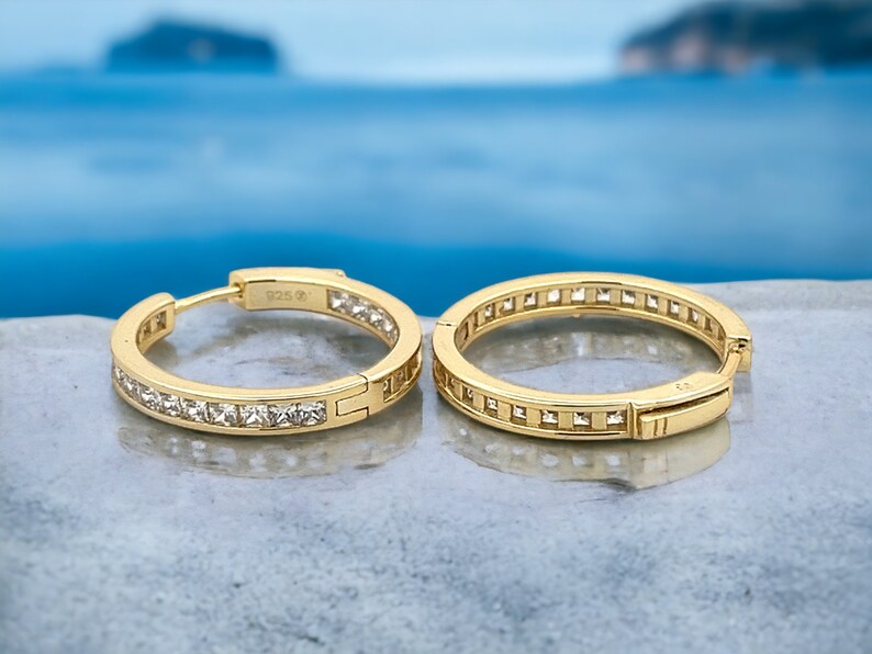 May include: A pair of gold hoop earrings with a row of clear cubic zirconia stones around the edge.