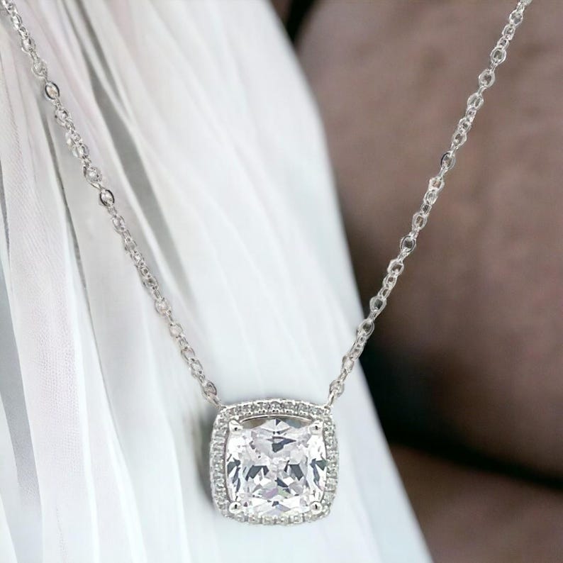 May include: A silver chain necklace with a large, square-shaped, clear cubic zirconia pendant. The pendant is surrounded by a halo of smaller cubic zirconia stones.
