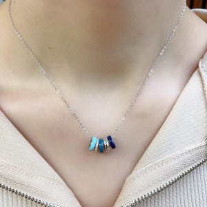 May include: A silver necklace featuring five colorful, cylindrical charms. The charms are in shades of turquoise, light blue, and dark blue, with one charm adorned with small, clear stones. The necklace is displayed against a neutral background.