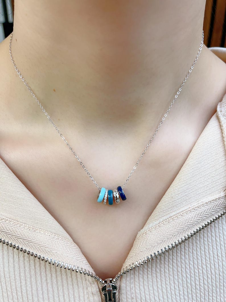 May include: A silver necklace featuring five colorful, cylindrical charms. The charms are in shades of turquoise, light blue, and dark blue, with one charm adorned with small, clear stones. The necklace is displayed against a neutral background.