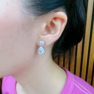 Pode incluir: Um par de brincos de prata em forma de l&aacute;grima com pedras de zirc&ocirc;nia c&uacute;bica transparentes. Os brincos t&ecirc;m uma pequena pedra redonda na parte superior e uma pedra maior em forma de l&aacute;grima pendurada abaixo.