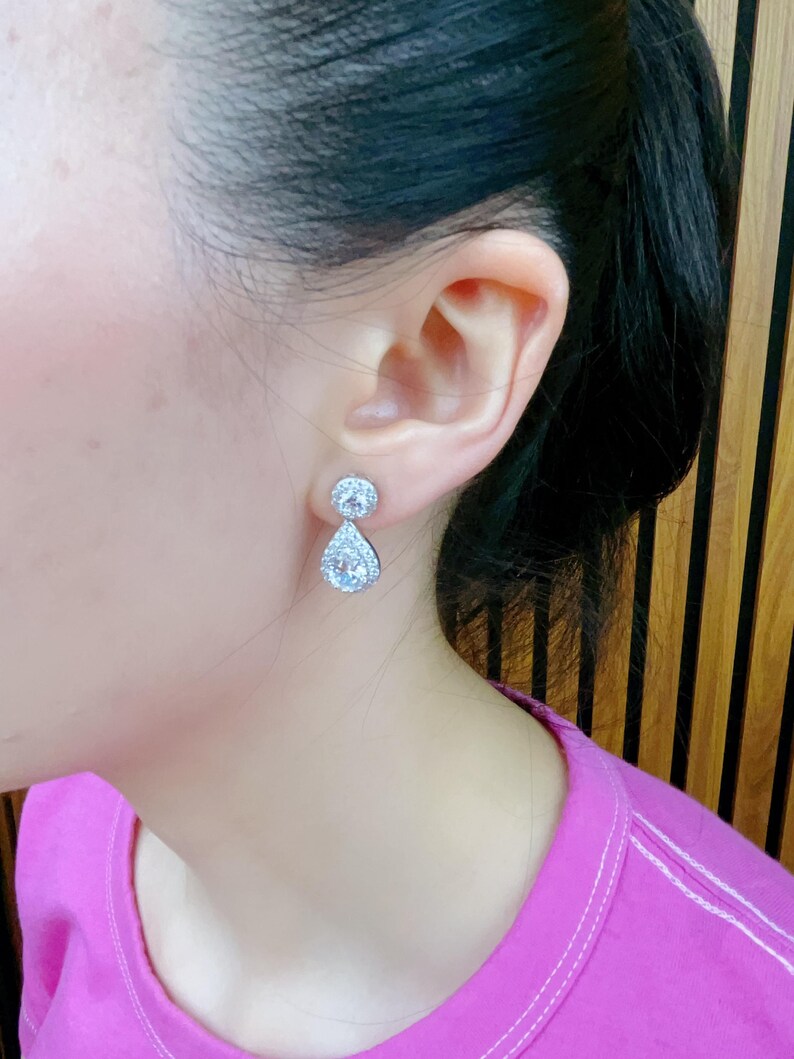 Pode incluir: Um par de brincos de prata em forma de l&aacute;grima com pedras de zirc&ocirc;nia c&uacute;bica transparentes. Os brincos t&ecirc;m uma pequena pedra redonda na parte superior e uma pedra maior em forma de l&aacute;grima pendurada abaixo.