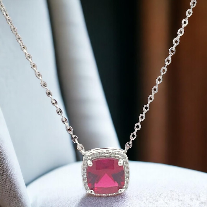 May include: A silver chain necklace with a square-shaped pendant featuring a large, bright red gemstone surrounded by smaller clear gemstones.