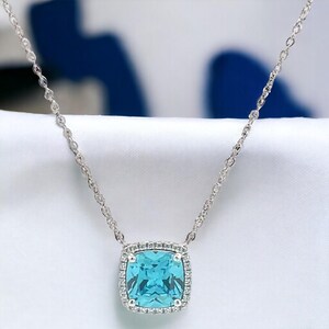 May include: A silver chain necklace with a square-shaped pendant featuring a large, light blue gemstone surrounded by a halo of smaller, clear gemstones.