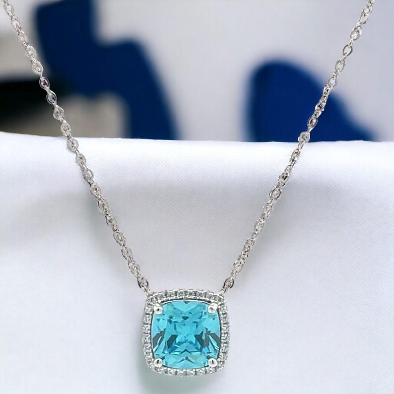 May include: A silver chain necklace with a square-shaped pendant featuring a large, light blue gemstone surrounded by a halo of smaller, clear gemstones.