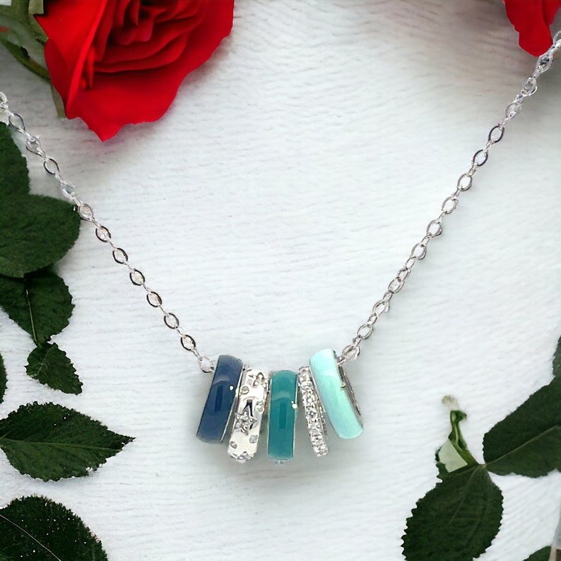 May include: A silver necklace featuring five rectangular pendants in shades of blue and silver. The pendants are arranged in a row and suspended from a delicate chain. Some pendants are adorned with rhinestones or star patterns. The necklace is displayed on a white surface.