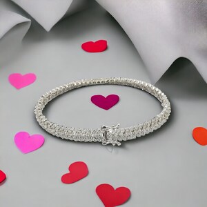 May include: A silver tennis bracelet with a row of sparkling cubic zirconia stones. The bracelet is on a gray background with pink hearts.