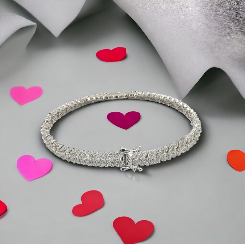 May include: A silver tennis bracelet with a row of sparkling cubic zirconia stones. The bracelet is on a gray background with pink hearts.