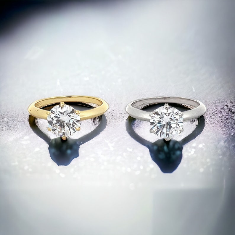 May include: Two engagement rings with a round brilliant cut diamond. The ring on the left is set in yellow gold, and the ring on the right is set in white gold.