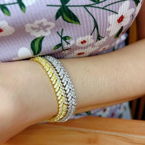 May include: A gold and silver tennis chain bracelet with a diamond-like finish. The bracelet is layered with three chains, each with a different color and style.