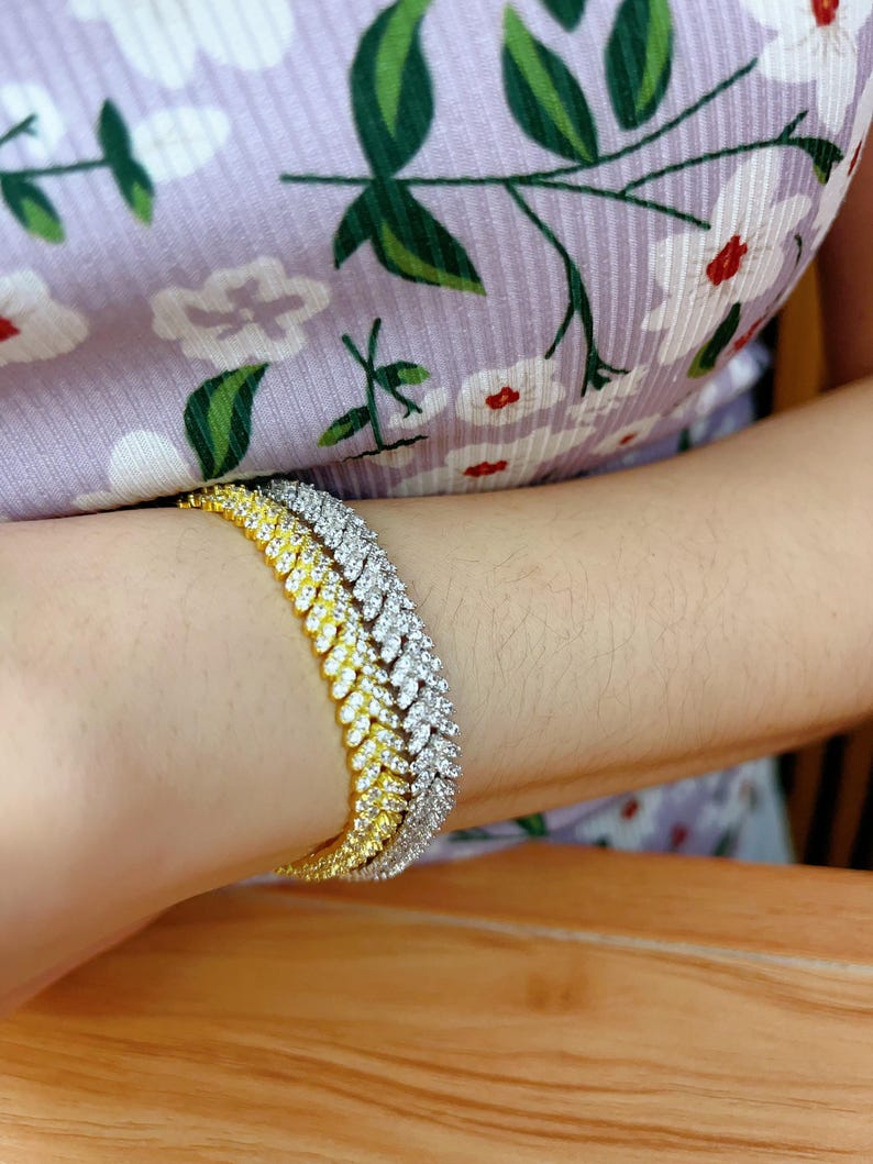 May include: A gold and silver tennis chain bracelet with a diamond-like finish. The bracelet is layered with three chains, each with a different color and style.