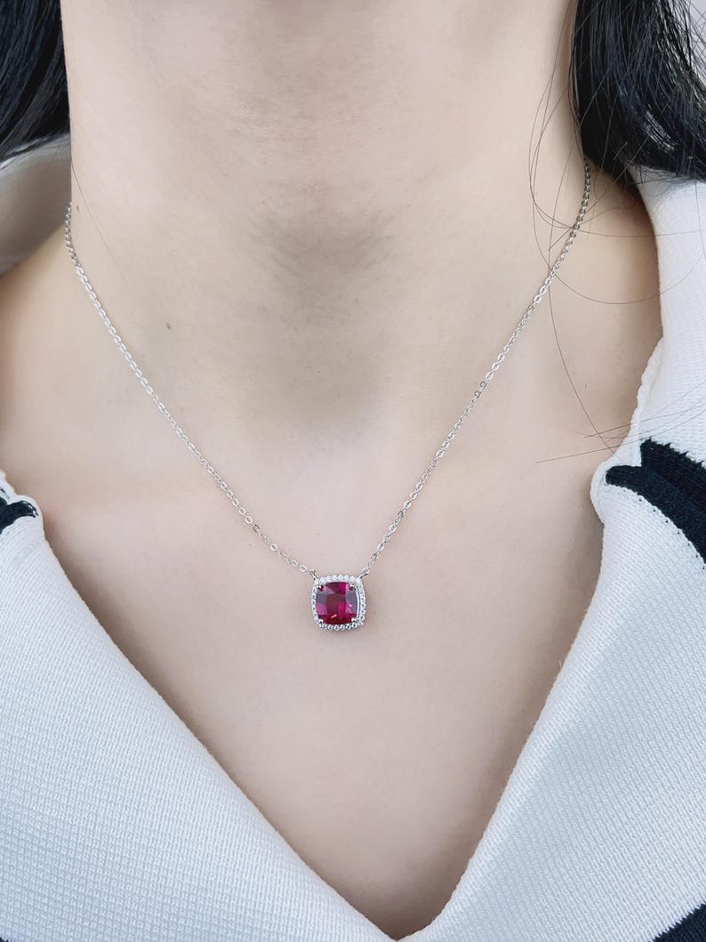 May include: A silver chain necklace with a red gemstone pendant. The pendant is surrounded by smaller clear gemstones.