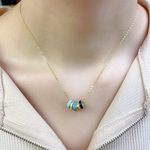 May include: A gold-colored necklace featuring a series of colorful enamel rings. The rings are in shades of blue, white, and gold. The necklace is worn around the neck and is displayed against a neutral background.
