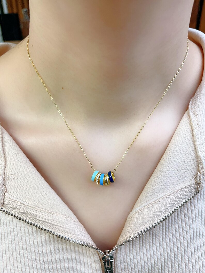 May include: A gold-colored necklace featuring a series of colorful enamel rings. The rings are in shades of blue, white, and gold. The necklace is worn around the neck and is displayed against a neutral background.