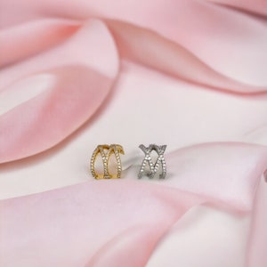 May include: Two ear cuffs with a criss-cross design, one is gold and the other is silver, both are set with clear cubic zirconia stones.