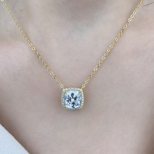 May include: A gold chain necklace with a square-shaped pendant featuring a large, clear, faceted gemstone surrounded by a halo of smaller, clear gemstones.
