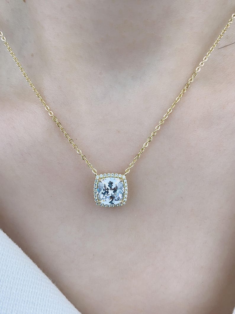 May include: A gold chain necklace with a square-shaped pendant featuring a large, clear, faceted gemstone surrounded by a halo of smaller, clear gemstones.