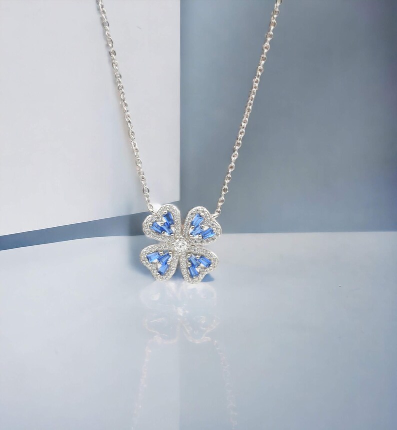 May include: A silver necklace with a blue and white flower pendant. The flower has four petals and is surrounded by small, clear stones.