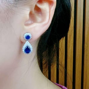 Pode incluir: Um par de brincos de prata com uma pedra preciosa azul em forma de l&aacute;grima cercada por pedras preciosas transparentes menores.