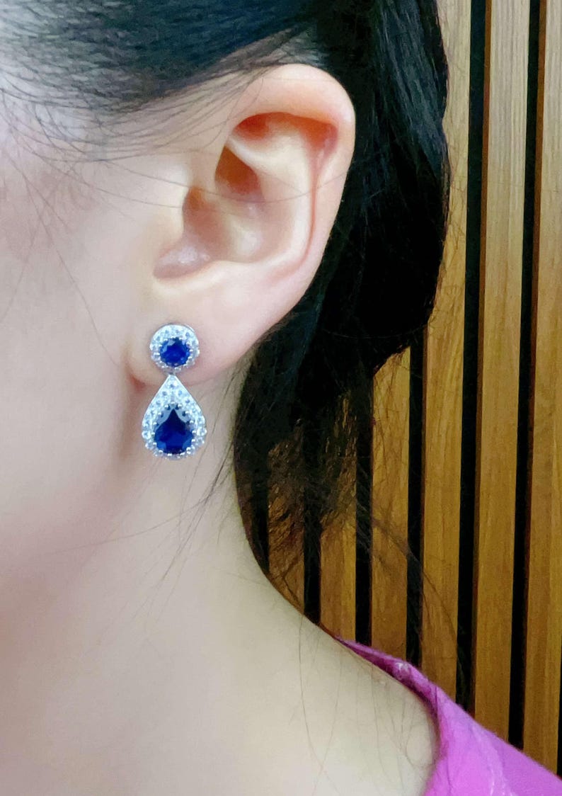 Pode incluir: Um par de brincos de prata com uma pedra preciosa azul em forma de l&aacute;grima cercada por pedras preciosas transparentes menores.
