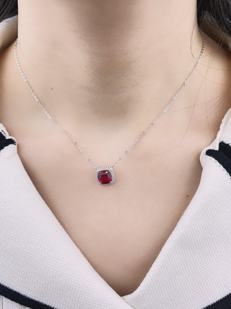 May include: A silver chain necklace with a red gemstone pendant. The pendant is surrounded by smaller clear gemstones.