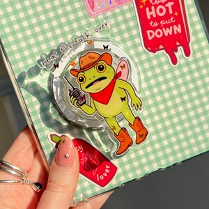 Op de afbeelding: Een doorzichtige iPad-hoes met een groen en wit ruitpatroon, versierd met stickers. Stickers zijn onder meer een roze schedel, een kikker met een cowboyhoed en de tekst "too HOT to put DOWN".