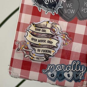 May include: A clear tablet case with a red and white gingham pattern. The case is decorated with several stickers, including a black heart with "Good Girl" and three gray heart-shaped stickers that say "I Vow, We Vow, You Vow". Other stickers include "Open Wider, Baby" and "Morally Grey".