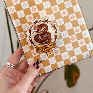 May include: Clear phone case with a yellow and white checkered pattern and small pink flowers. A sticker with a brown snake, barbed wire, and the words "Stay Venomous" is centered on the case.