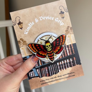 May include: A circular device grip featuring a moth with orange and red wings and a skull-shaped body. The moth is centered on a white background with small gold stars. The packaging card displays the text Kindle & Device Grip and a library scene.