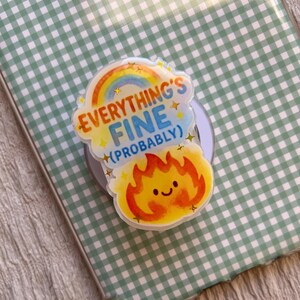 May include: A phone grip with the text "EVERYTHING'S FINE (PROBABLY)", a rainbow, and a smiling flame graphic. The grip is on a phone case with a green and white gingham pattern.