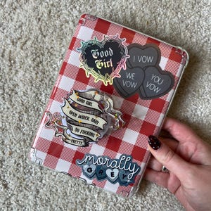 May include: A clear phone case with a red and white gingham pattern, adorned with various stickers. The stickers include phrases like "Good Girl," "I Vow," and "Morally Grey." The case is held by a person.