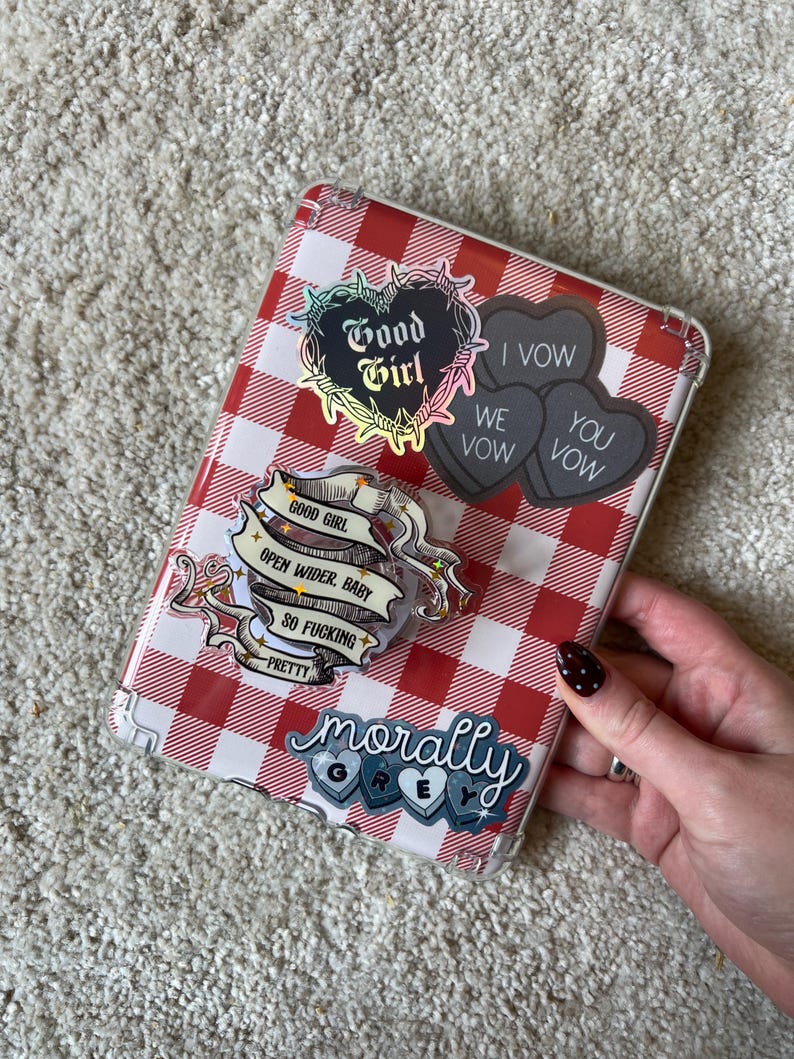 May include: A clear phone case with a red and white gingham pattern, adorned with various stickers. The stickers include phrases like "Good Girl," "I Vow," and "Morally Grey." The case is held by a person.