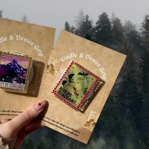 Puede incluir: Dos accesorios Kindle & Device Grip. Uno presenta una escena de monta&ntilde;a con el texto "City of Starlight", el otro el texto "Riders Quadrant" y una ilustraci&oacute;n de drag&oacute;n. Presentados sobre cartulina beige, con un fondo de bosque.