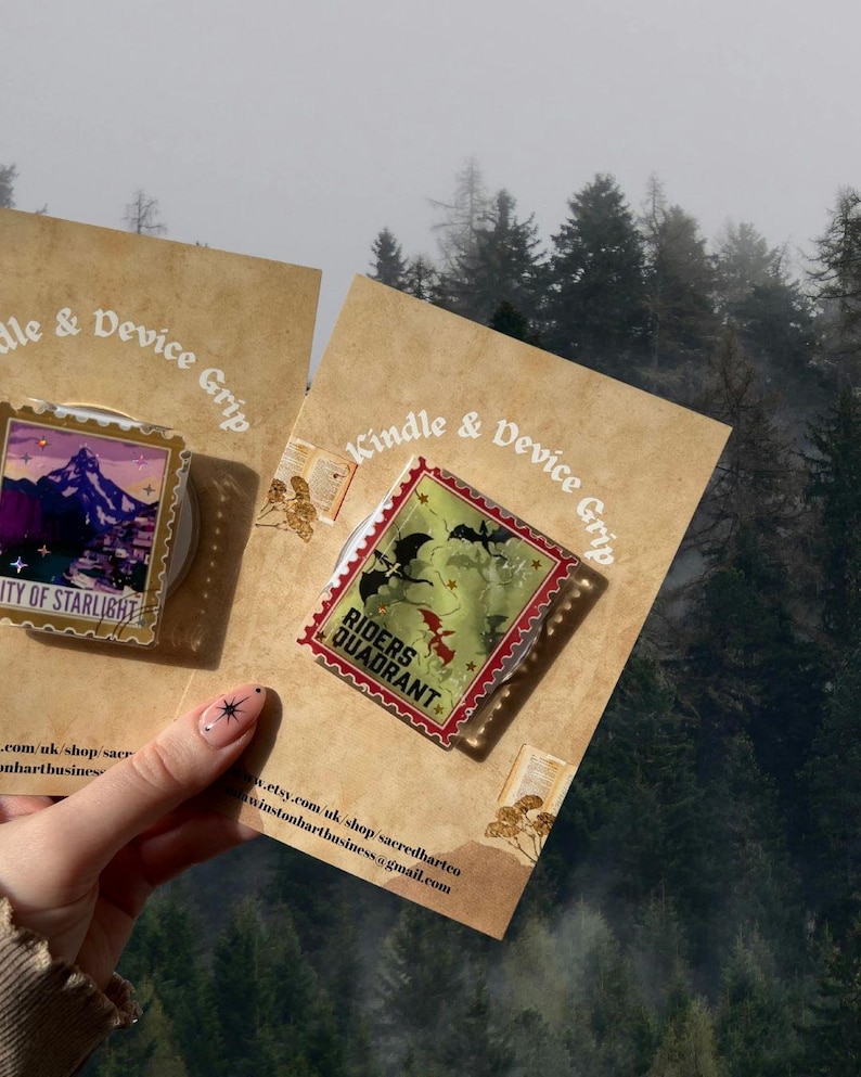 Puede incluir: Dos accesorios Kindle & Device Grip. Uno presenta una escena de monta&ntilde;a con el texto "City of Starlight", el otro el texto "Riders Quadrant" y una ilustraci&oacute;n de drag&oacute;n. Presentados sobre cartulina beige, con un fondo de bosque.