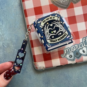 May include: A clear phone case with a red and white checkered pattern. A blue book-shaped keychain with the text "YOU HAD ME AT TRIGGER WARNINGS" and a skeleton hand design. A matching charm with heart cutouts and star accents.