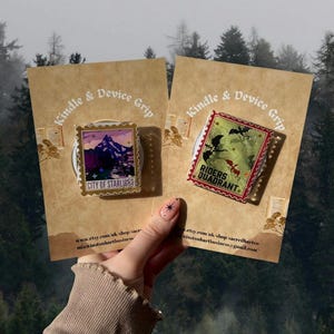 Puede incluir: Dos agarres rectangulares para Kindle y dispositivos en tarjetas marrones. Uno presenta una escena de monta&ntilde;a con el texto "CITY OF STARLIGHT". El otro tiene un dise&ntilde;o verde y rojo con "RIDERS QUADRANT". Ambos tienen un dise&ntilde;o de sello postal.