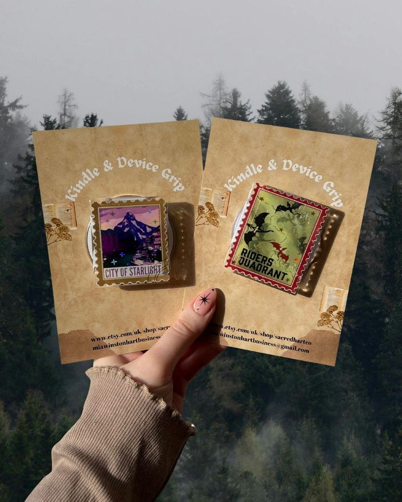 Puede incluir: Dos agarres rectangulares para Kindle y dispositivos en tarjetas marrones. Uno presenta una escena de monta&ntilde;a con el texto "CITY OF STARLIGHT". El otro tiene un dise&ntilde;o verde y rojo con "RIDERS QUADRANT". Ambos tienen un dise&ntilde;o de sello postal.