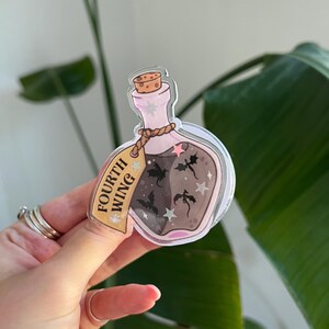 May include: A clear, bottle-shaped sticker with a cork top and a label that reads "FOURTH WING." The bottle contains a dark liquid with silhouettes of dragons and stars. The sticker has a pink and gold color scheme.