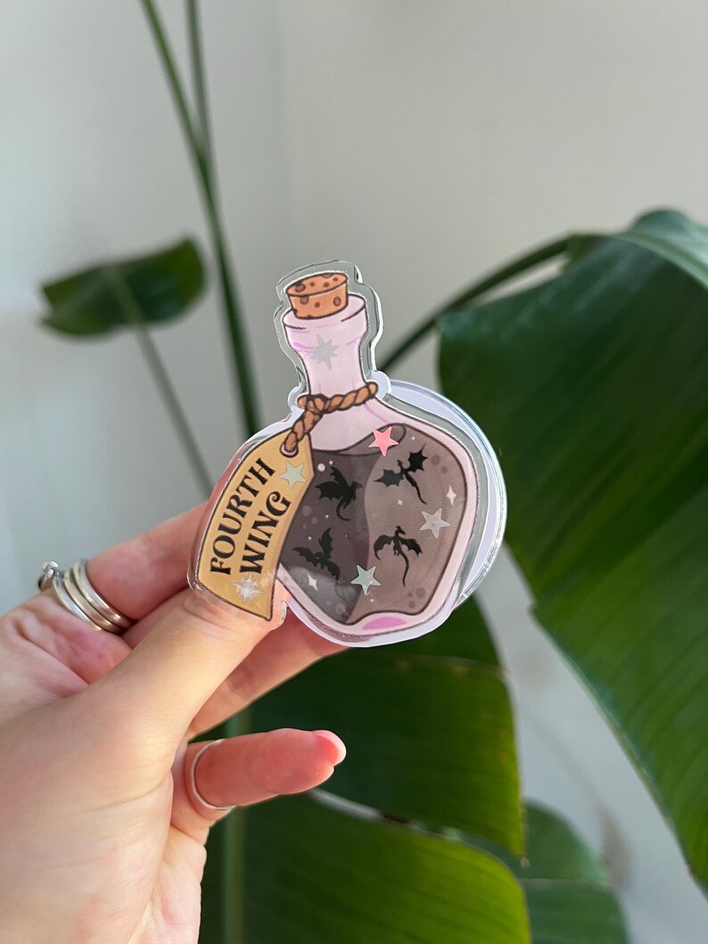May include: A clear, bottle-shaped sticker with a cork top and a label that reads "FOURTH WING." The bottle contains a dark liquid with silhouettes of dragons and stars. The sticker has a pink and gold color scheme.
