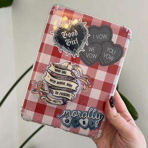 May include: A clear tablet case with a red and white checkered pattern. The case is decorated with stickers that read "Good Girl", "I Vow, We Vow, You Vow", "Open Wider, Baby, So Fucking Pretty", and "Morally Grey".