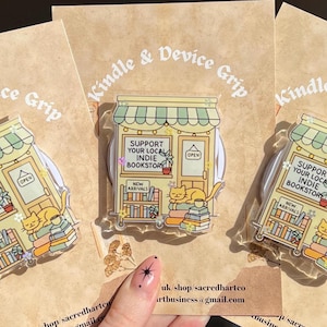 May include: Clear acrylic Kindle and device grip featuring a yellow indie bookstore design. The design includes a cat, books, and the text "Support Your Local Indie Bookstore" and "New Arrivals". The grip is on a brown card.