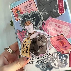 May include: A clear phone case covered in stickers. Stickers include a potion bottle with the words "Fourth Wing", a heart-shaped sticker that says "I Vow", and a pink ticket that says "Try Crime".