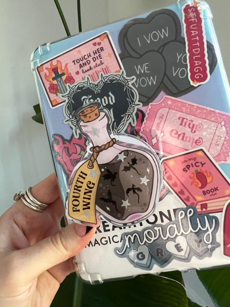 May include: A clear phone case covered in stickers. Stickers include a potion bottle with the words "Fourth Wing", a heart-shaped sticker that says "I Vow", and a pink ticket that says "Try Crime".