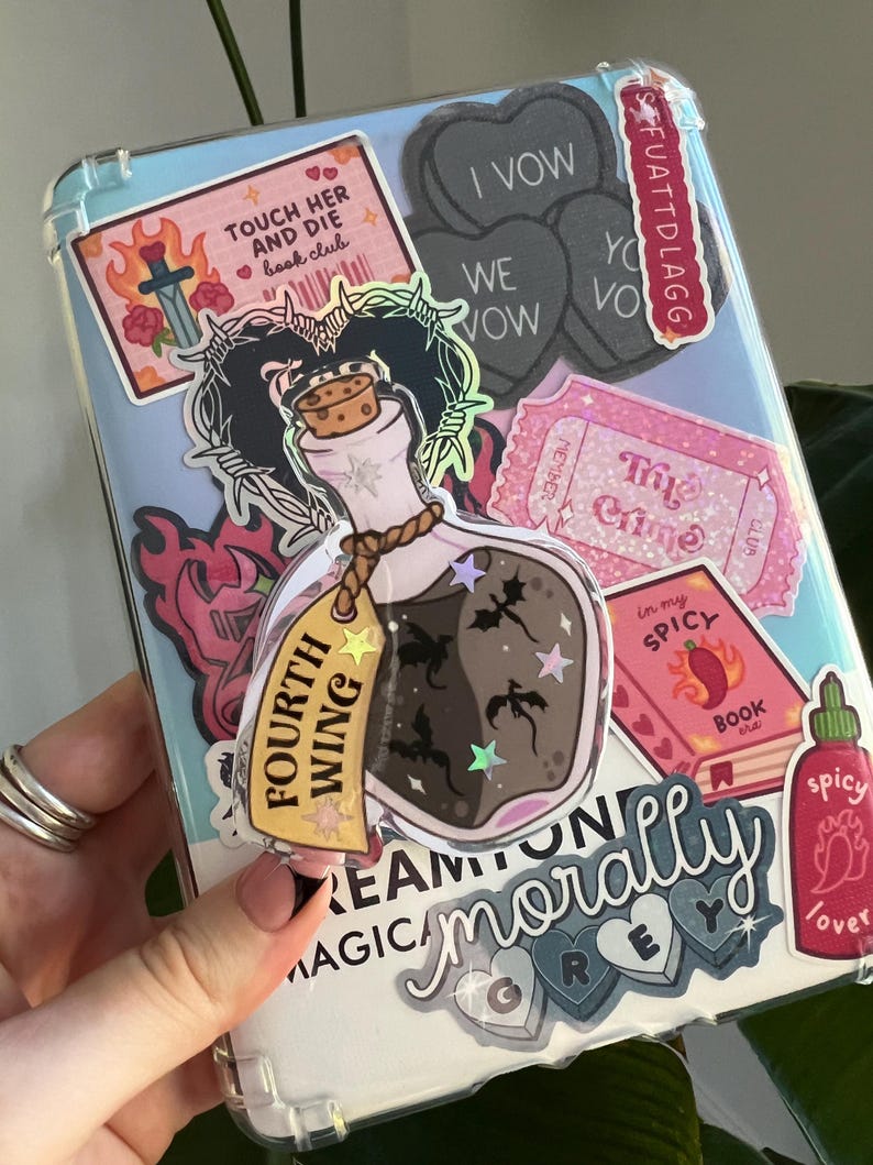 May include: A clear phone case covered in stickers. Stickers include a potion bottle with the text "Fourth Wing", a pink book with "Spicy Book Club", and heart-shaped stickers with "I Vow" and "We Vow".