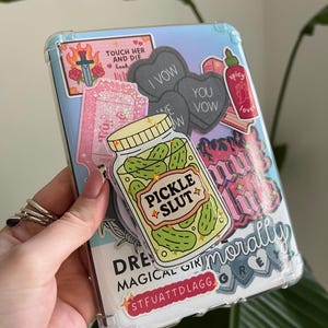 Puede incluir: Funda transparente para teléfono decorada con varias pegatinas. Las pegatinas incluyen un tarro de pepinillos con "Pickle Slut", pegatinas en forma de corazón con "I Vow" y "You Vow", y una pegatina que dice "Touch Her and Die book".
