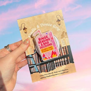 May include: A pink and white Kindle & Device Grip with the text "Binge Reading & Bad Decisions" and "Regret-Free Reading Fuel". The grip is shaped like a juice box with flames at the bottom. The background is a library scene.