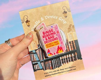 Könnte beinhalten: Ein pinkfarbener und weißer Kindle & Device Grip mit dem Text "Binge Reading & Bad Decisions" und "Regret-Free Reading Fuel". Der Grip hat die Form eines Saftkartons mit Flammen am Boden. Der Hintergrund ist eine Bibliotheksszene.