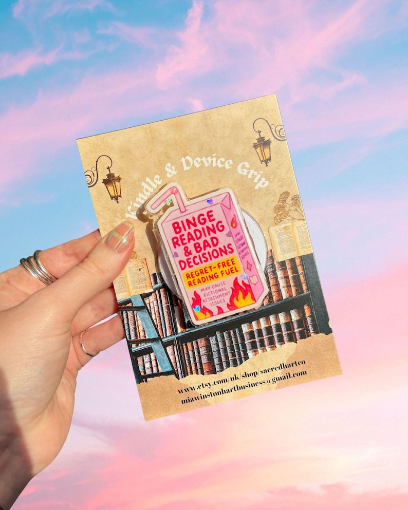 May include: A pink and white Kindle & Device Grip with the text "Binge Reading & Bad Decisions" and "Regret-Free Reading Fuel". The grip is shaped like a juice box with flames at the bottom. The background is a library scene.