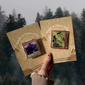 Puede incluir: Dos pines en tarjetas marrones con el texto "Kindle & Device Grip". Un pin presenta una escena de monta&ntilde;a con "CITY OF STARLIGHT". El otro pin tiene un fondo verde con "RIDERS QUADRANT".