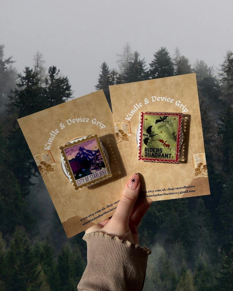 Puede incluir: Dos pines en tarjetas marrones con el texto "Kindle & Device Grip". Un pin presenta una escena de monta&ntilde;a con "CITY OF STARLIGHT". El otro pin tiene un fondo verde con "RIDERS QUADRANT".